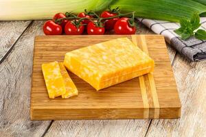 A piece of cheese on a cutting board next to a tomato and a bunch of other vegetables photo