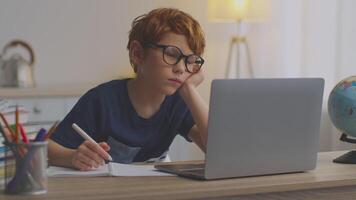 A red-haired boy sits at his desk, focused on a laptop screen while studying. He appears engaged in a lesson, surrounded by school supplies and a globe, in a well-lit room. video