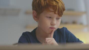 A young boy with ginger hair focuses intently on his online lesson while studying at home. He appears deep in thought, raised finger indicating concentration during learning. video