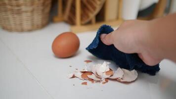 Hand cleaning up broken eggshells with a blue cloth on a white countertop, a whole brown egg and wicker basket blurred in the background video