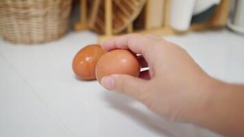 Close-up of hands cracking a brown egg on a white countertop, with wicker basket and blurred kitchen items in the background video