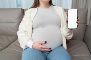 Close-up of pregnant woman holding and showing mobile phone with white empty display at home. Mockup image with copy space for app or interface customization. E-commerce. photo