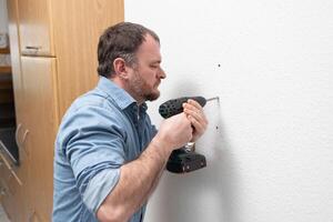 drilling a hole in wall. man drills hole in wall using a drill. drill helps to do this work neatly and accurately. holes can be used to screw in screws as a mount for pictures, shelves, etc. photo