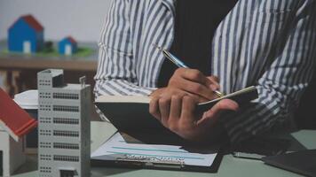 A woman writing in a notebook with a pen and a house model in the background video