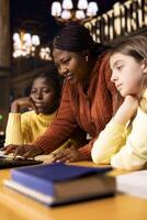 African american private tutor offering support to two schoolgirls, guiding them through a lesson plan at a library desk. Teacher providing learning materials, mentoring scholars. photo