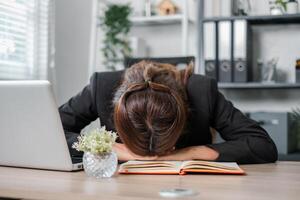 Tired businesswoman in office resting her head on a desk with a laptop and notebook, symbolizing stress and burnout. photo