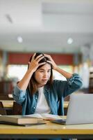 Young woman in a classroom, looking frustrated with her laptop, hands on head, surrounded by books. photo