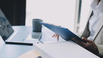 A woman in a business suit is holding a clipboard and looking at a laptop video