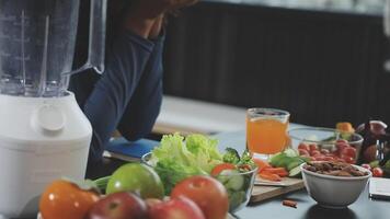 A woman sitting at a table with a blender and a bowl of fruit and vegetables video