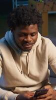 Vertical African American man on a sofa texting mates in his living room, entertained by social media and messages. Depiction of remote work breaks, digital communication and relaxation at home. Camera B. video
