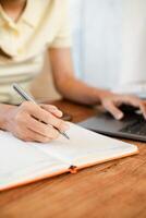 Close-up of a person writing in a notebook with a pen while using a laptop, showcasing productivity and focus. photo
