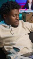 Vertical Young adult working remotely from his loft apartment on a laptop. Black freelancer focusing on tasks, blending productivity and relaxation while browsing the web from a cozy sofa. Camera B. video