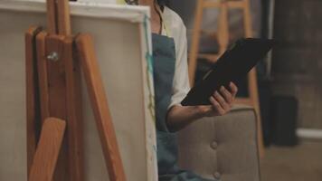 A woman is holding a tablet while sitting in front of an easel video