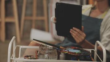 A woman is holding a tablet computer in front of a table with paint supplies video