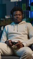 Vertical Cheerful man in his apartment playing games on couch, enjoying day off from work. Black gamer battling enemies in online multiplayer shooter on gaming console. Camera A. video