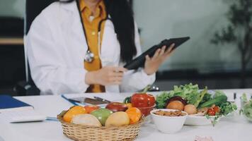 A woman in a white coat is holding a tablet while sitting at a table with fruit and vegetables video