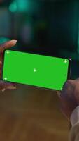 Vertical African american person using device with green screen from the comfort of his modern trendy home, sitting on the couch and working in the living room. Happy confident young man. Camera A. video