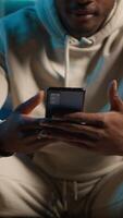 Vertical Relaxed black freelancer chatting with friends on his smartphone via texts, sitting on the couch. Young adult showcasing laughter and leisure, staying entertained while working remotely. Camera A. video