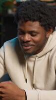 Vertical African American man on a sofa texting mates in his living room, entertained by social media and messages. Depiction of remote work breaks, digital communication and relaxation at home. Camera A. video