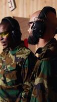 Vertical Portrait of joyous army soldiers wearing safety gear in shooting gallery, preparing to fire at targets. Smiling military units in uniform and protection equipment testing accuracy, camera B video