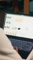Vertical Remote worker accountant doing math calculations using financial graphs on laptop, working at home. Self employed guy in loft comparing business economic figures on notebook. Camera A. video