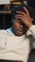 Vertical Self employed man suffering from pain and high fever sitting on sofa, trying to work and finish his tasks. Black male feelancer suffering from illness and feeling sick, using laptop. Camera B. video