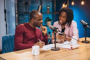 Two African American podcasters are recording a new episode, discussing and taking notes while consulting a smartphone photo