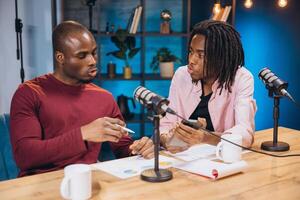 African American podcasters discussing data charts, recording episode with smartphone and professional microphone in studio setting photo