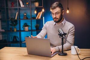 Podcaster speaking into condenser microphone while working on laptop in home recording space photo