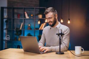 Young man working in a cozy home studio, recording a podcast while using a laptop and microphone, enjoying a warm beverage photo