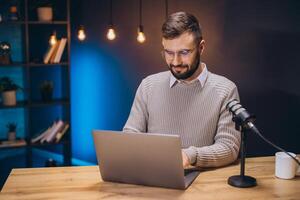 Young podcaster preparing content using laptop with microphone in home studio with blue light and bookcase photo