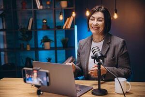 Smiling female podcaster recording audio content, speaking into microphone while using laptop and smartphone in home workspace photo
