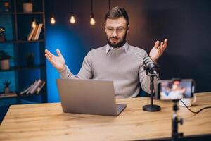 Podcaster working at home studio, speaking into microphone, laptop and smartphone nearby, using hands while talking photo