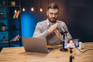 Young man gesturing while recording a podcast and live streaming using professional microphone and smartphone in home studio photo