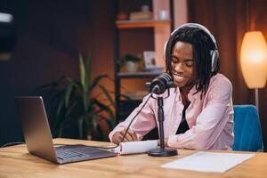 African American man content creator recording a podcast episode in a cozy home studio, smiling while taking notes and engaging with the microphone photo