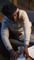 Vertical Casual person sitting On the sofa and reading research papers with analytics, multitasking with laptop at home. Man diving into professional or personal topics of improvement, independent. Camera B. video