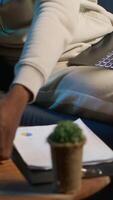 Vertical Casual person sitting On the sofa and reading research papers with analytics, multitasking with laptop at home. Man diving into professional or personal topics of improvement, independent. Camera A. video