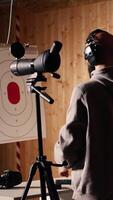 Vertical Firing range customer looking in zooming device to check if he shot bullseye next to instructor overseeing client. Person analyzing targets shot using magnifying lens during firearm practice, camera A video