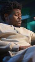 Vertical Self employed man suffering from pain and high fever sitting on sofa, trying to work and finish his tasks. Black male feelancer suffering from illness and feeling sick, using laptop. Camera A. video