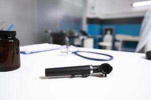 Selective focus of otoscope placed on desk essential medical equipment for ear examination. In empty office white table having doctor tools and bottle of pills ready for use at patient appointment. photo