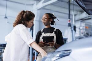Seasoned mechanic next to client in car service orders new engine for damaged vehicle using tablet. Employee looking for components on internet to replace old ones in defective automobile photo