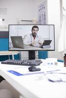 Desktop in vacant physician office displays consultation screen. Computer monitor captures doctor providing medical advice through virtual call emphasizing use of technology in patient care. photo