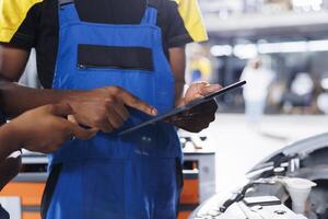 African american engineer trainee uses tablet in car service to watch tutorial on how to replace motor. Garage workspace intern looks up training videos online on device, unsure how to fix vehicle photo