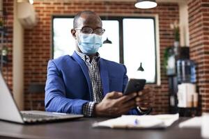 Portrait of businessman with face mask texting on his mobile device in brick wall office. African american manager taking a break at work, using smartphone for chatting and checking social media. photo
