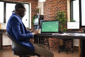 African american employee sitting at office desk and looking at laptop with greenscreen. Businessman working at table as digital device displays blank mockup template in brick wall workspace. photo