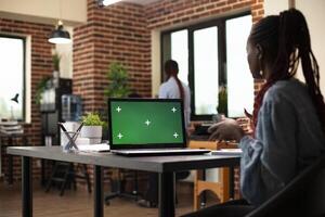 Female manager seated at desk having an online discussion as laptop displays isolated chromakey template. Digital device with greenscreen is set on table while black woman does a virtual presentation. photo