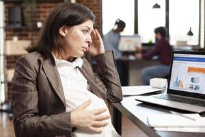 Closeup of exhausted pregnant woman resting her head in hand, doing market analysis on laptop in busy startup office. Tired expectant mother touching her baby bump and reviewing data charts on device. photo