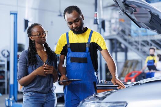 BIPOC mechanic in car service uses tablet to calculate invoice for client after repairing motor. African american employee in garage with customer calculating final costs after servicing vehicle photo