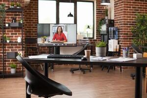 Modern office with desktop pc monitor on clean table displaying virtual session. Shelves with file folders and plants arranged in spacious room, along with videocall of businesswoman on computer. photo