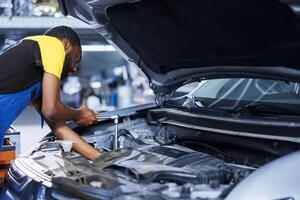 BIPOC mechanic in car service uses torque wrench to tighten nuts after replacing damaged engine. Hardworking garage employee uses professional tools to fix customer automobile photo